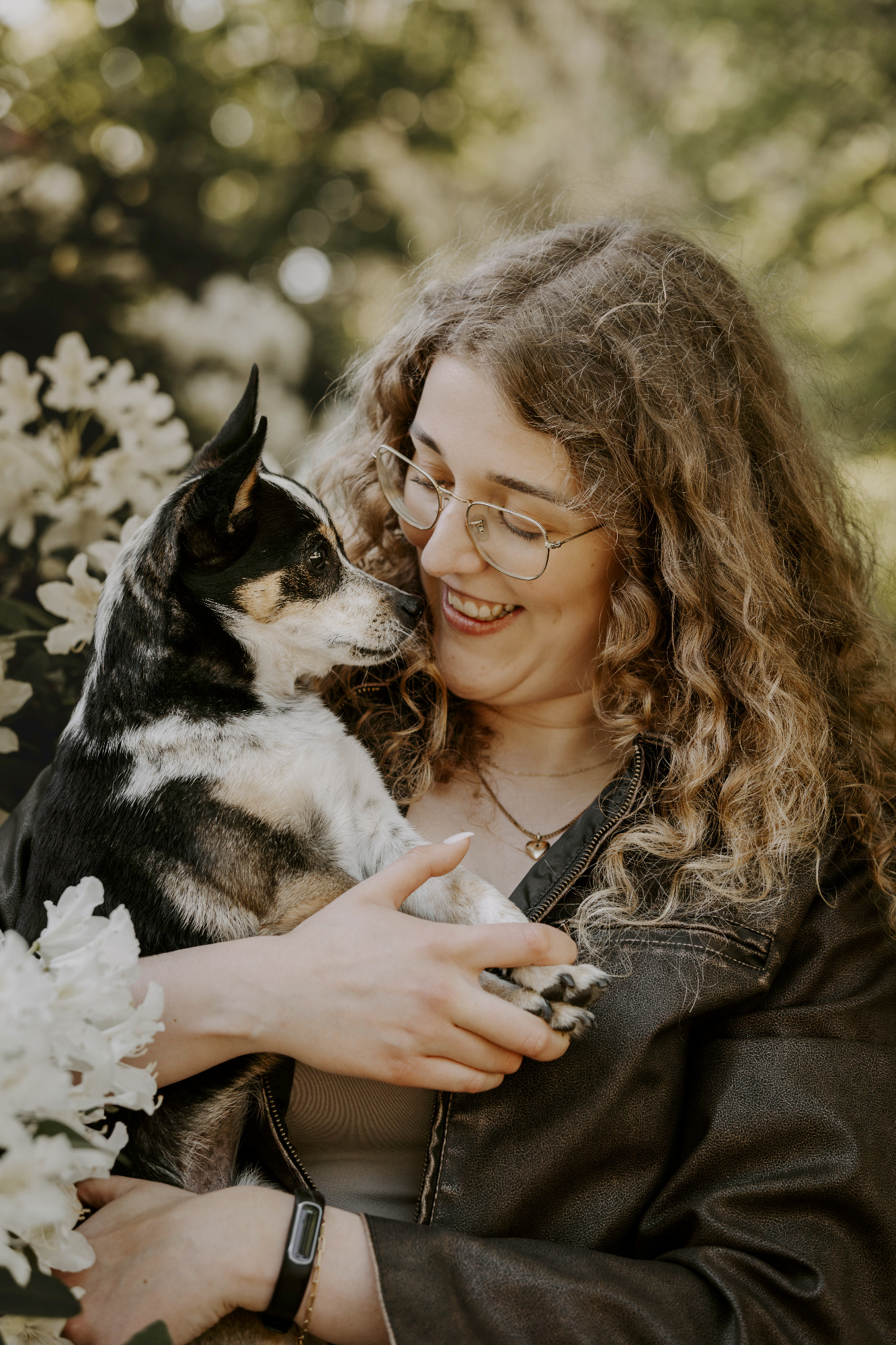 Eine Person mit lockigem Haar hält einen Hund und lächelt glücklich. Blumen im Hintergrund.